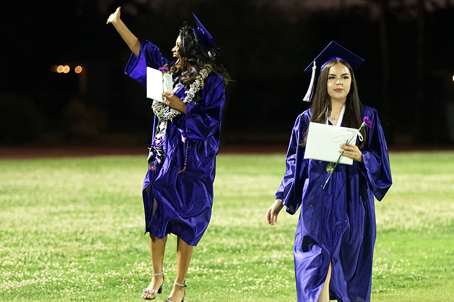 A graduate waves to her family as she and another graduate walk across the field