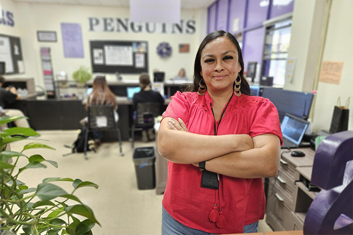 A woman in a red shirt smiles with her arms crossed in the school's front office