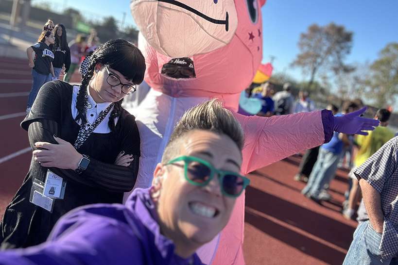 A woman with short hair and sunglasses smiles in front of a girl in a Wednesday Addams costume and a person in a pink inflatable unicorn costume