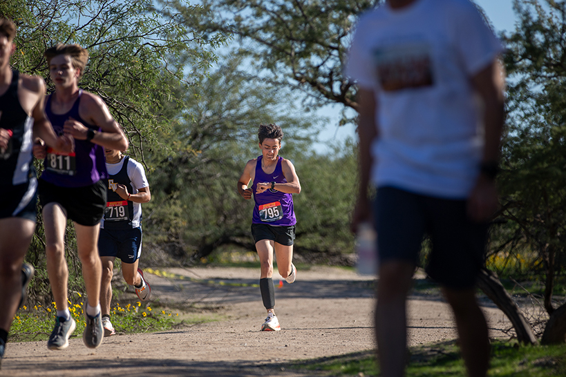 A teen boy looks down at his watch as he runs through the park behind other teen boys