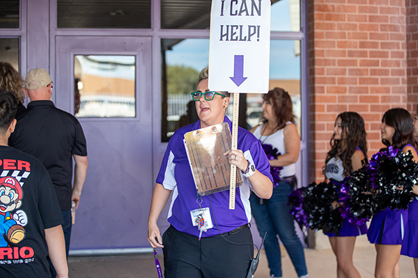 A woman in green glasses and a purple shirt holds up a sign reading I Can Help!
